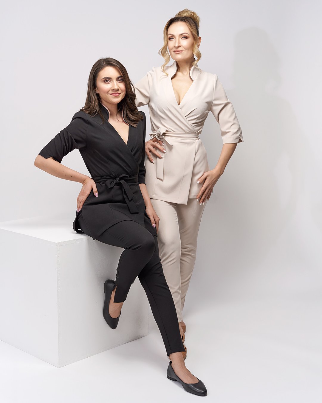 Two women, one seated on a white block, the other standing, dressed in professional attire, posing against a plain white background.