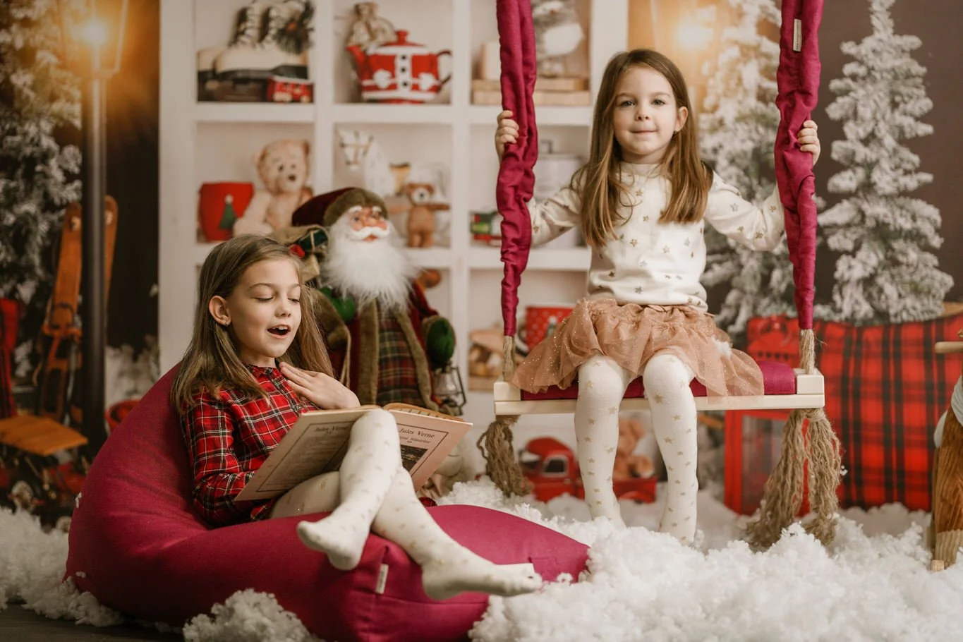 Two young girls with long brown hair, one sitting on a pink beanbag and the other on a swing, surrounded by Christmas decorations like decorated trees, ornaments, and stuffed animals, in a cozy holiday setting.