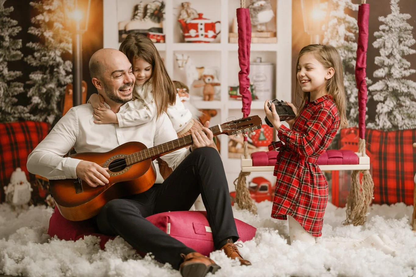 A man playing guitar and two young girls, one hugging him and the other holding a camera, in a decorated Christmas room with snow, trees, and festive decorations.