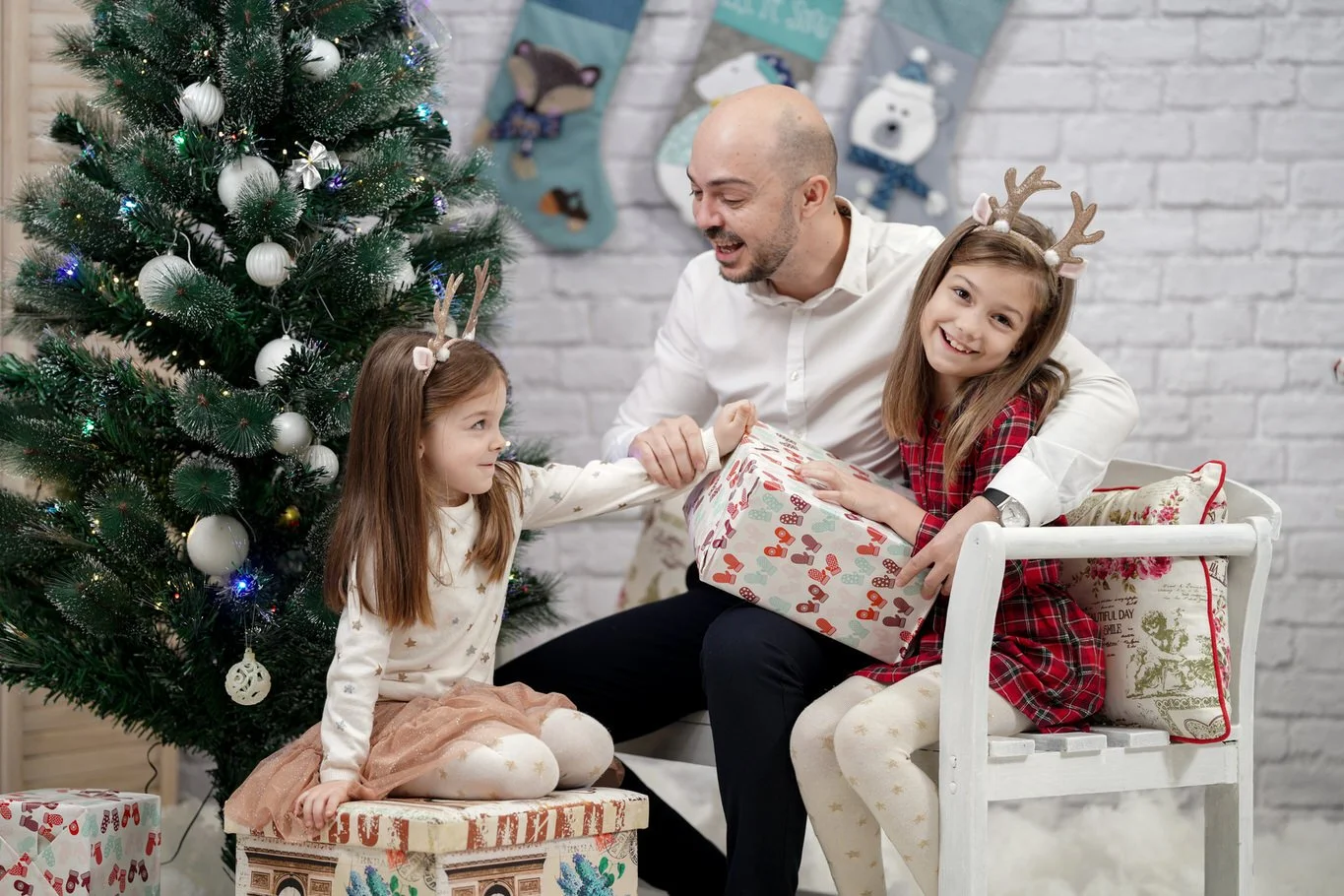 A father and two young girls exchanging Christmas presents near a decorated Christmas tree, all wearing reindeer antler headbands, in a festive living room.