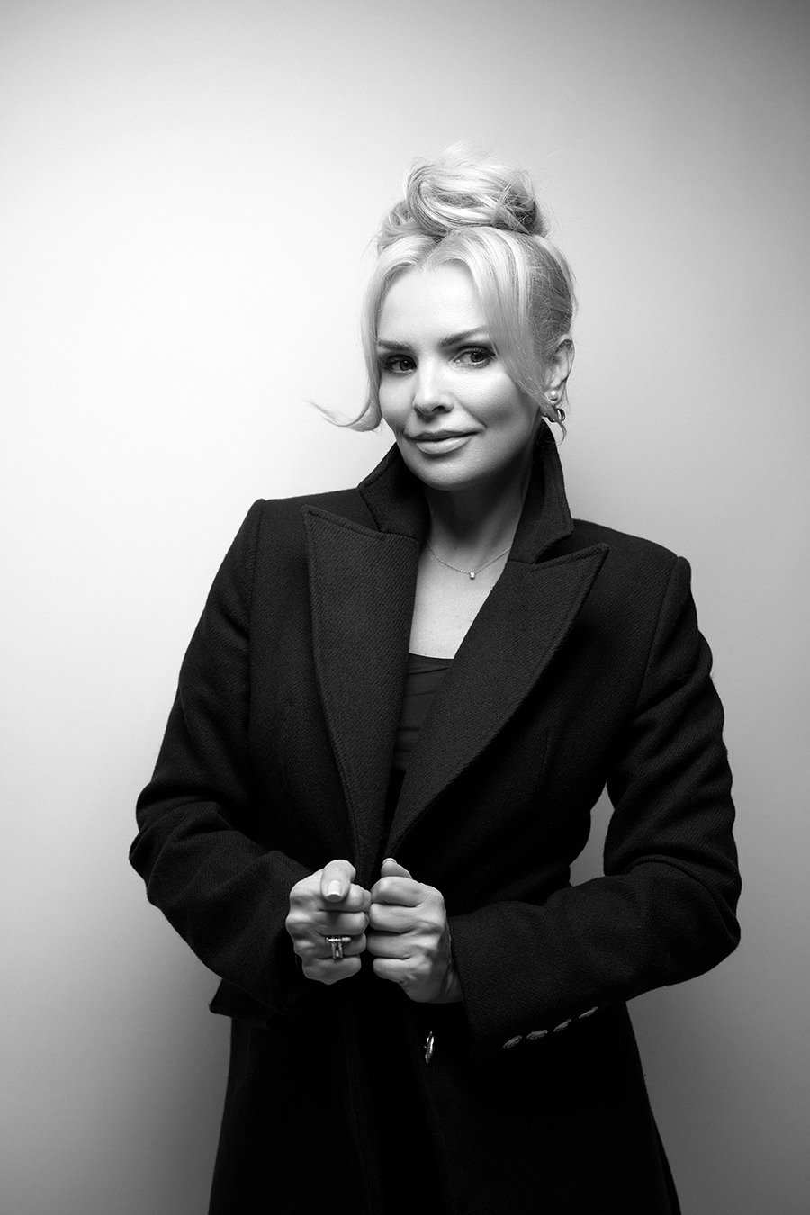 Black and white photo of a woman with styled blonde hair, wearing a dark coat, standing against a plain background, smiling subtly at the camera.