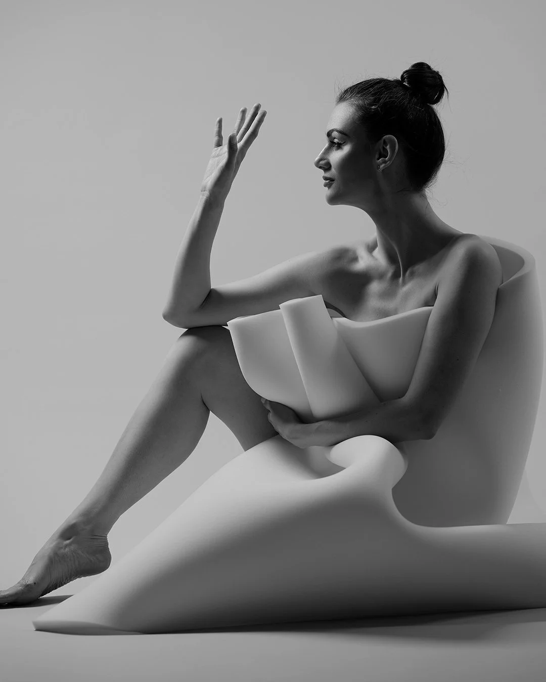 Black and white photo of a woman sitting on a modern, abstract chair, looking to her left with her hand raised near her face, with her dark hair styled in a top knot.
