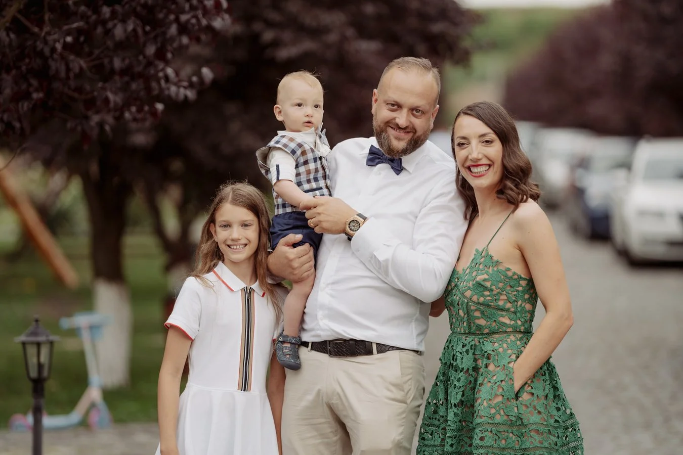 A family of four smiling outdoors, standing on a pathway with trees and parked cars in the background. The father is holding a young boy, and a woman and girl are standing beside them.