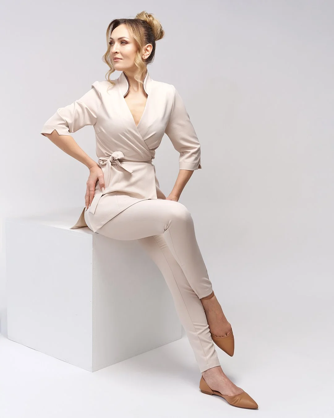 A woman with blonde hair styled in loose waves, wearing a light beige wrap dress with 3/4 length sleeves, sitting on a white block against a plain white background.