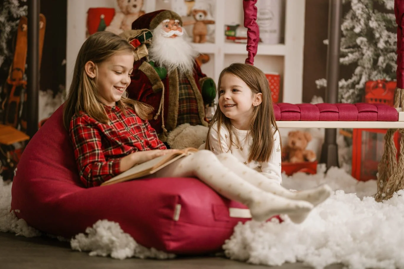 Two young girls sitting on a red bean bag, smiling and reading a book together in a festive Christmas setting with a decorated tree and teddy bears.