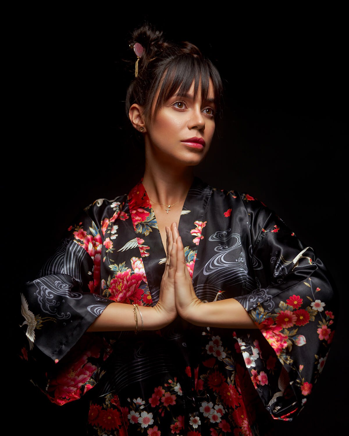 A woman in a black floral kimono with her hands together in a prayer-like pose, against a black background.