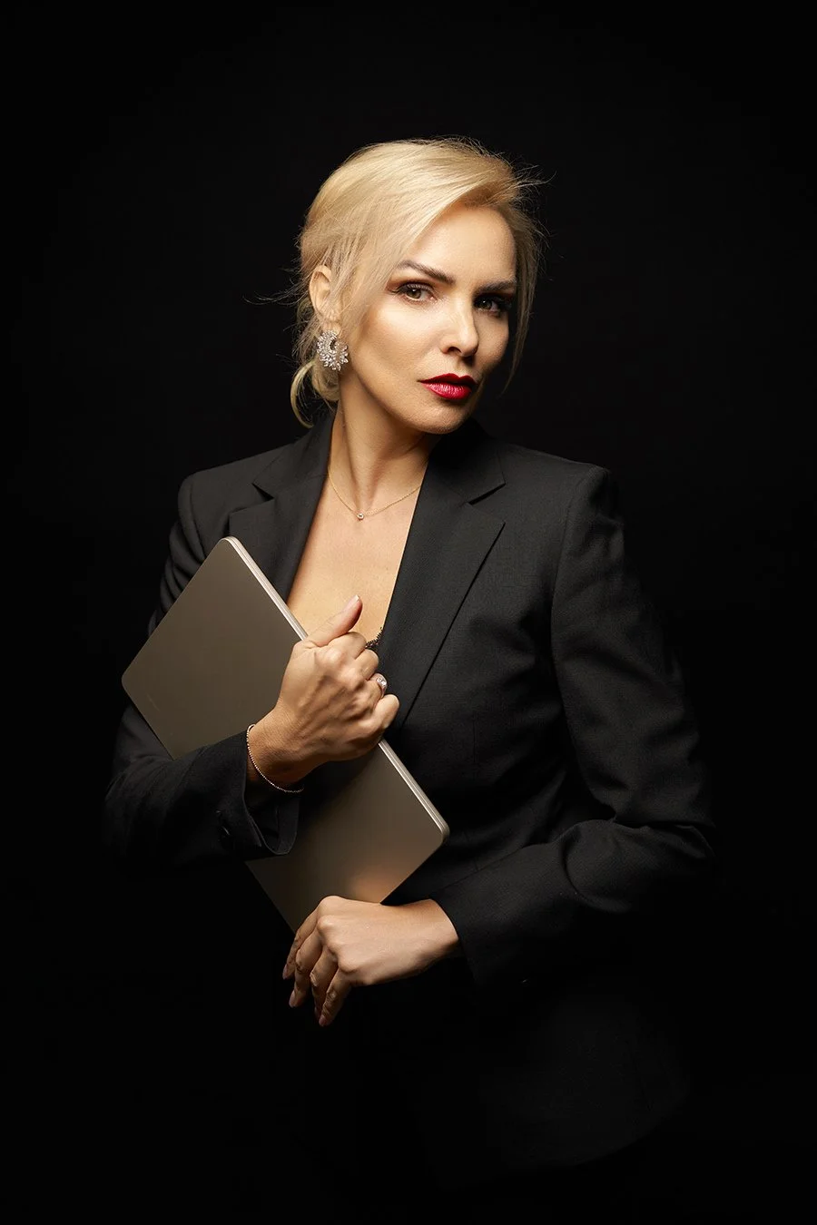 A blonde woman in a black blazer holding a closed silver laptop against a black background.