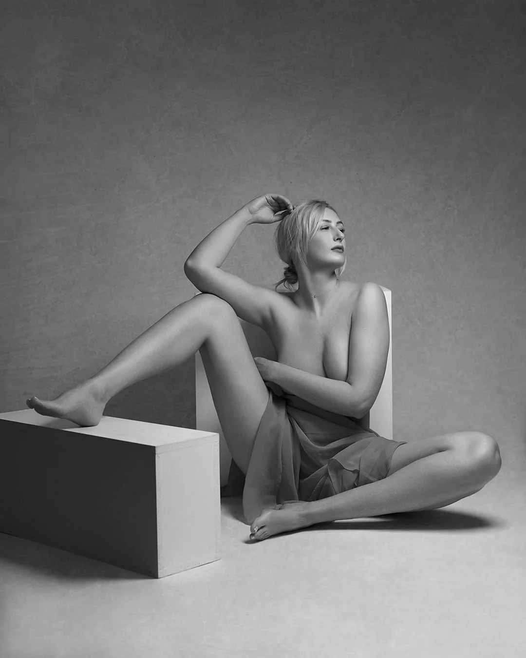 A black and white photograph of a woman sitting on the floor, partially covered by a sheer fabric. She has short blonde hair, and is posing with one arm behind her head and the other resting on her foot. Her legs are crossed, with one foot on a recta