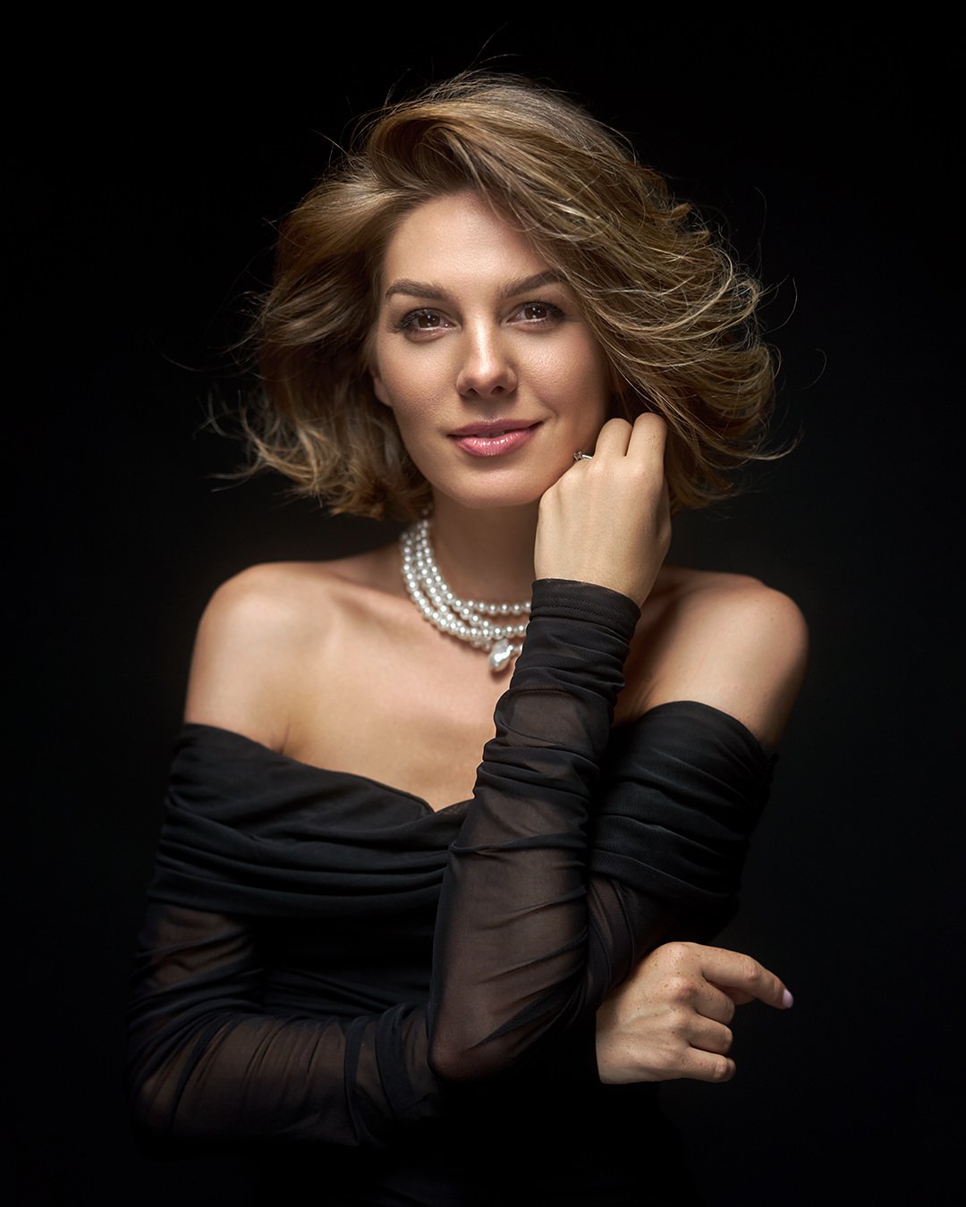Portrait of a woman with short wavy hair, wearing a black off-shoulder dress, a pearl necklace, and long black gloves, against a black background.