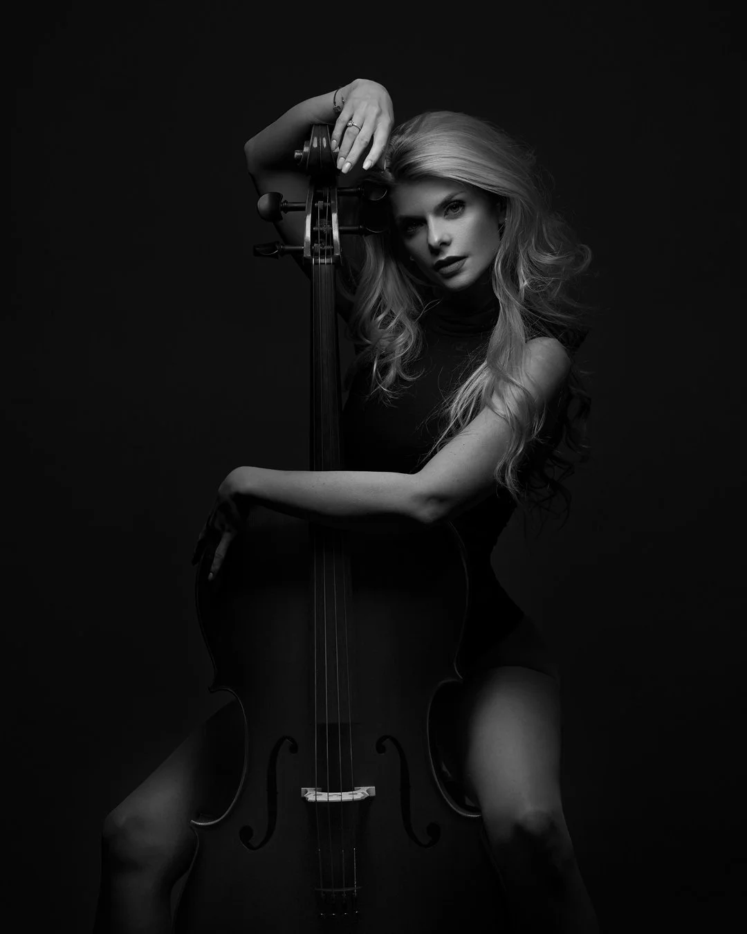 A woman with long, wavy hair poses with a cello, resting her right arm on the instrument's neck and holding the top of the cello with her left hand, against a black background.