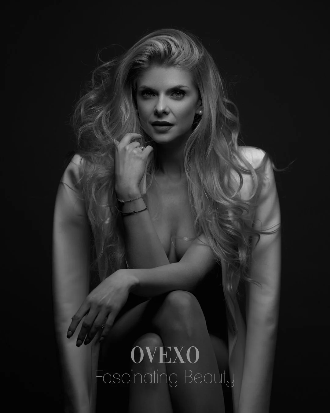 Black and white photo of a woman with long, wavy hair, seated with her hand near her face, wearing a blazer and jewelry, with the text 'OVEXO Fascinating Beauty' at the bottom.