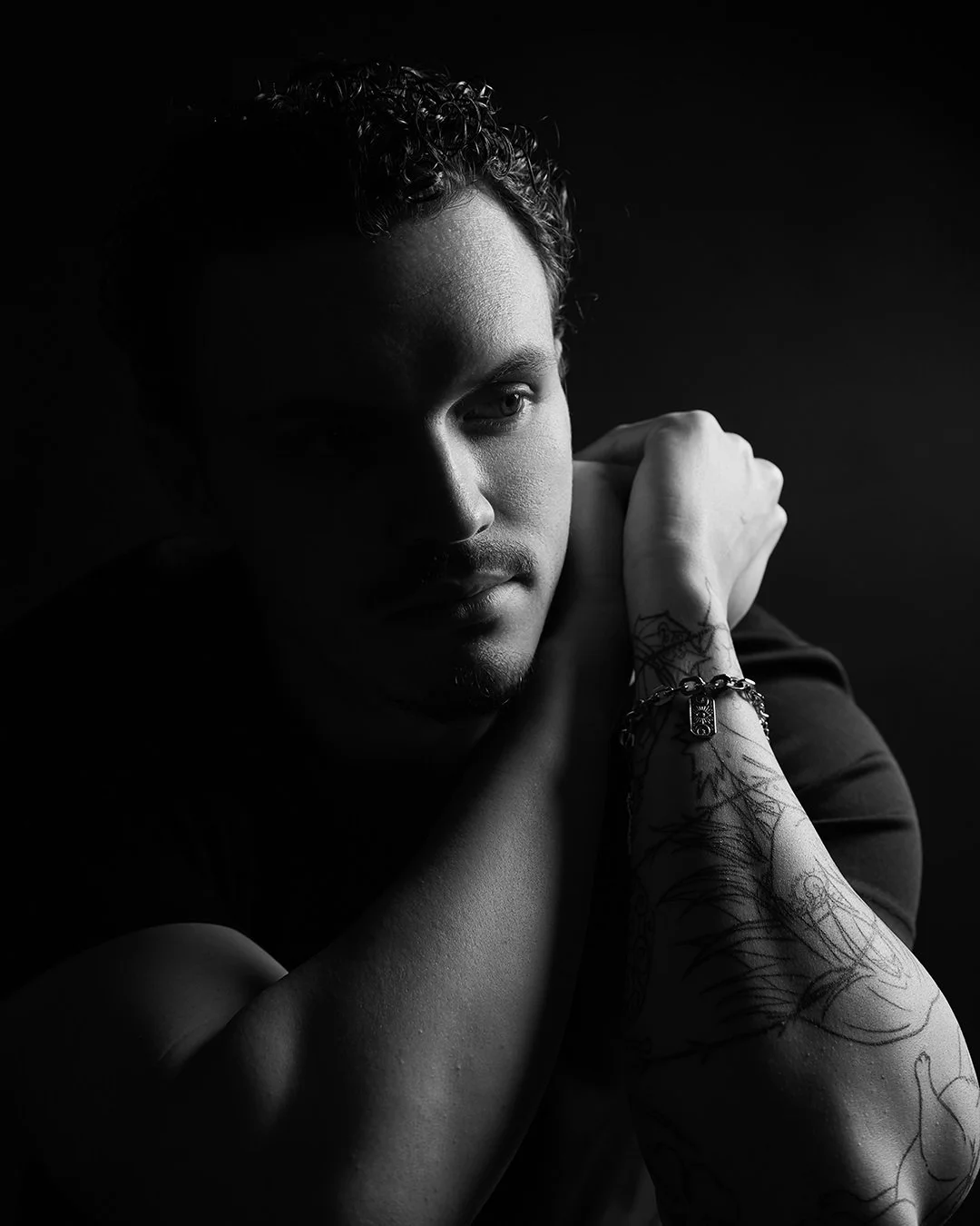 Un portret alb-negru al unui tânăr cu părul creț, cu bărbia sprijinită pe mână, cu tatuaje și o brățară pe braț, pe un fundal întunecat.