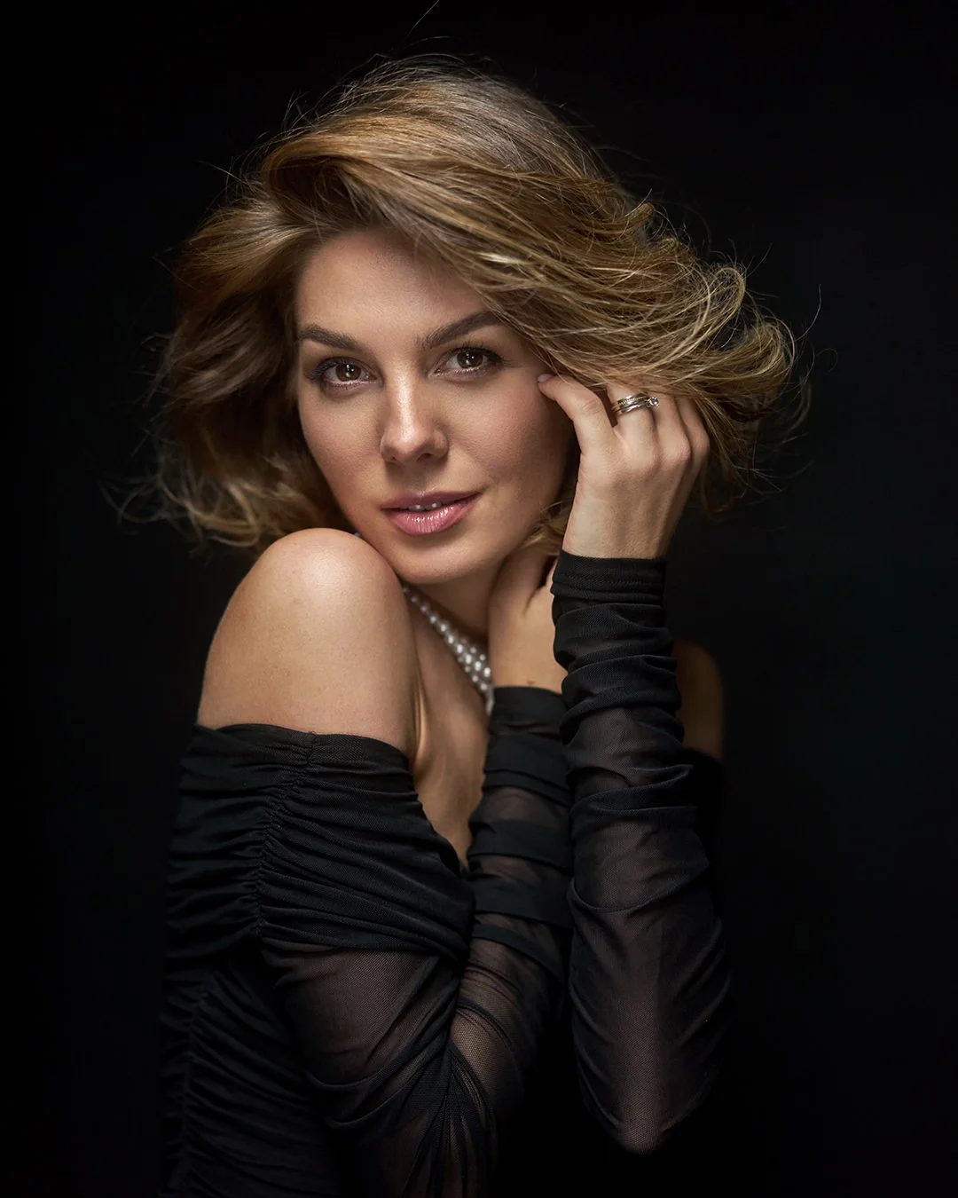 A woman with shoulder-length wavy hair wearing a black off-shoulder dress and black gloves, posed against a dark background.