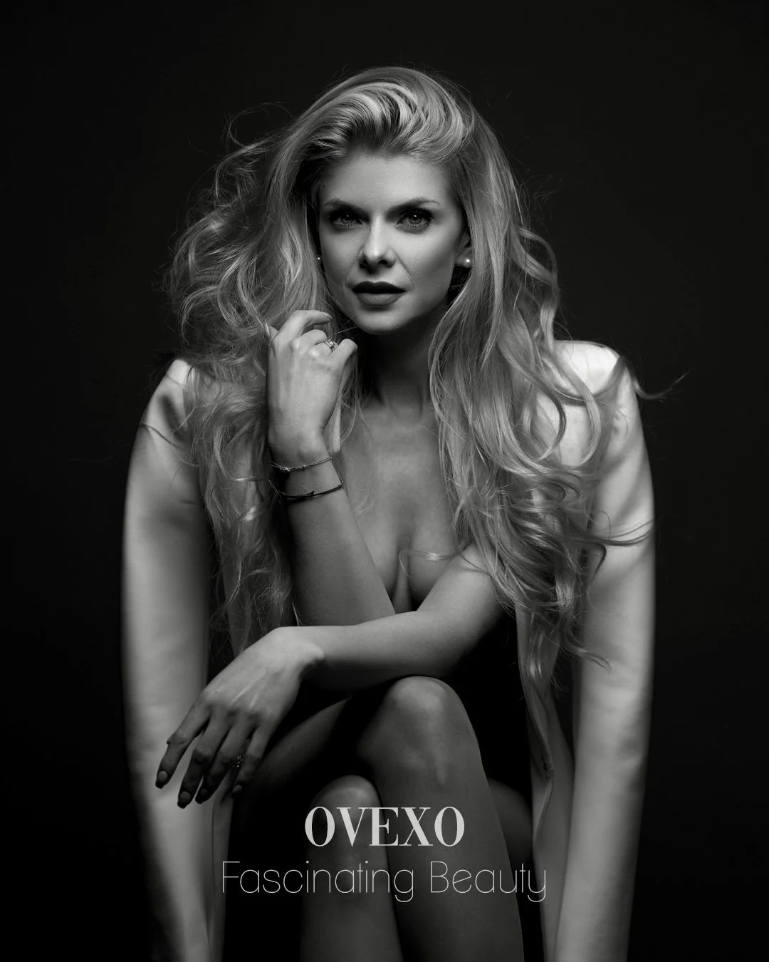 Black and white portrait of a woman with long, wavy hair, looking directly at the camera. She is wearing jewelry and is posed with one hand near her face. The text 'OVEXO Fascinating Beauty' is at the bottom of the image.