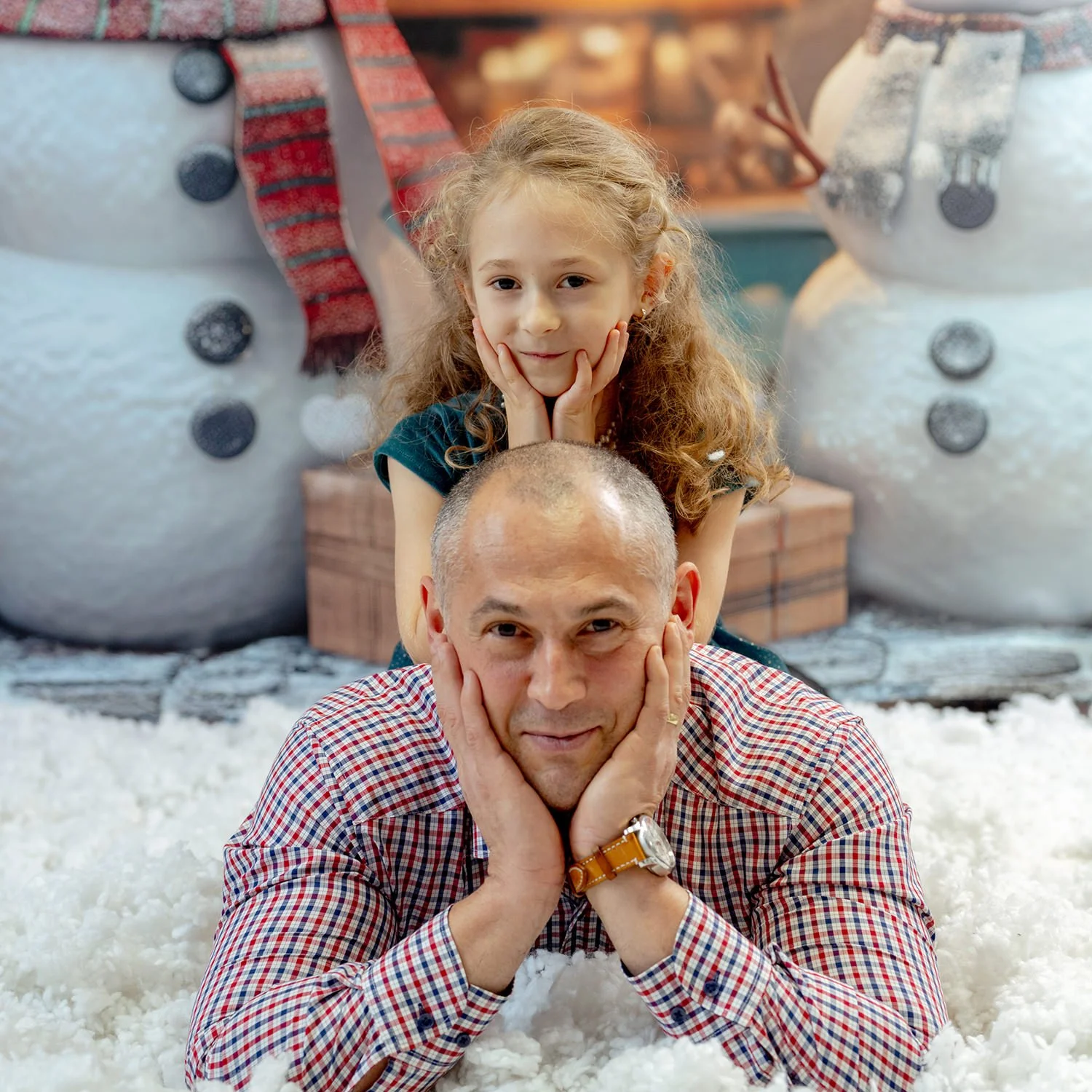 A man and a young girl are lying on a fluffy white surface, with the girl resting her chin on her hands and the man resting his chin on his hands, both smiling gently at the camera. Behind them are large snowman decorations with black buttons and a red-checkered scarf, creating a festive winter scene.