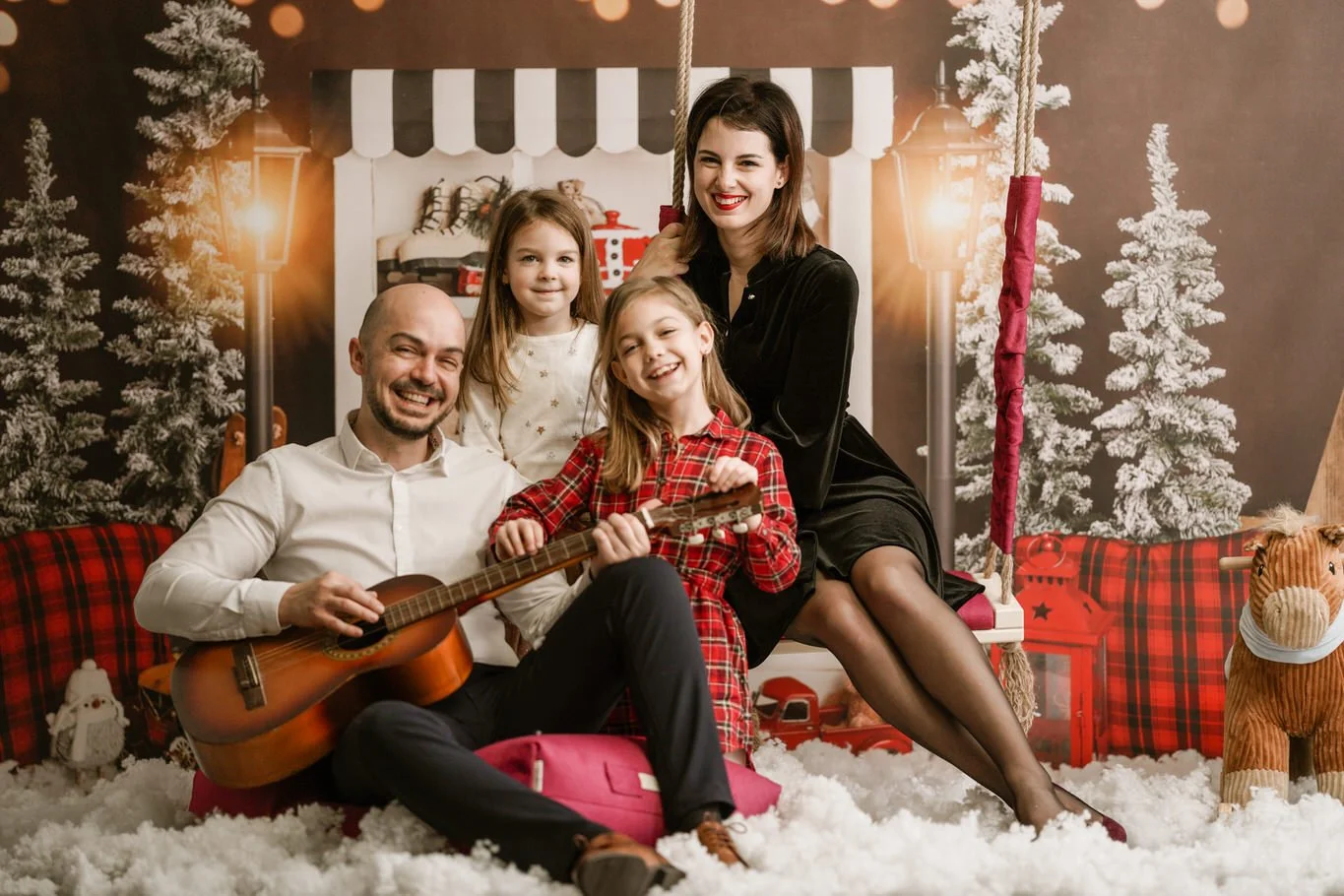 A family of four with two young girls and two adults sitting on a Christmas-themed swing surrounded by holiday decorations, including snow, Christmas trees, and stuffed animals, with everyone smiling and enjoying a festive moment.