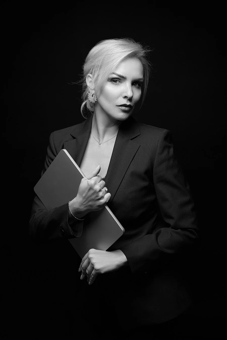 Black and white portrait of a woman with blonde hair, wearing a blazer, holding a closed laptop, looking confidently at the camera against a dark background.