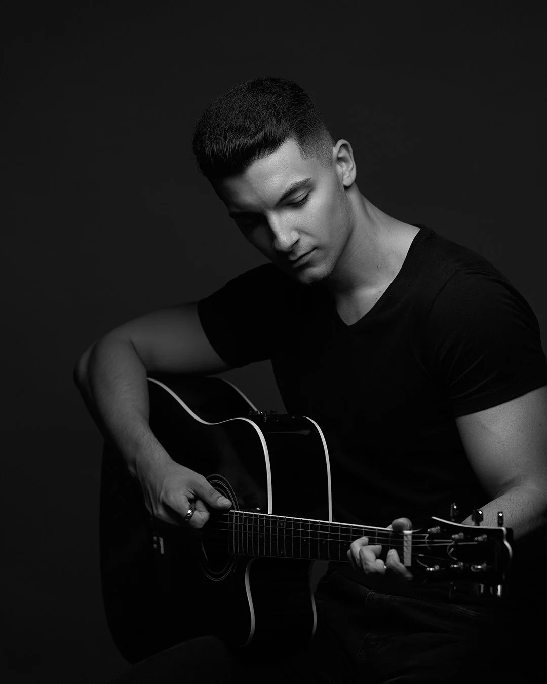 A young man with short hair playing an acoustic guitar against a dark background.