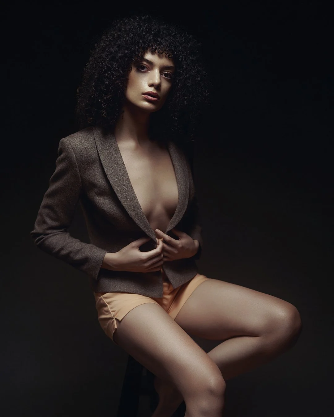 A woman with dark curly hair wearing a brown blazer and beige shorts against a black background.
