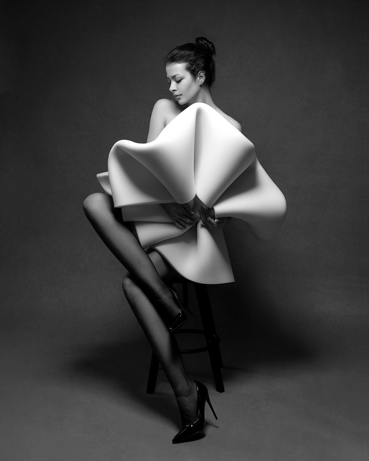 A black and white photo of a woman sitting on a stool, wearing a large, structured dress with an abstract, sculptural design, and black high heels. She has her hair tied up and is looking down with a calm expression.