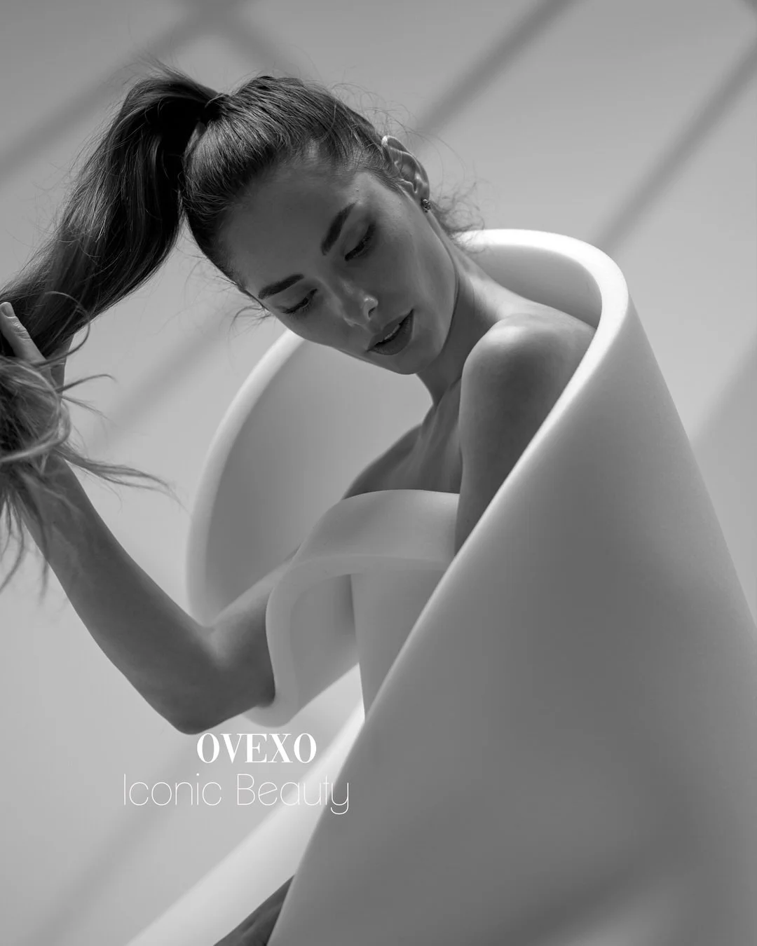 A black and white photo of a woman with a high ponytail, relaxed in a modern, sculptural bathtub, with the text "OVEXO Iconic Beauty" at the bottom left corner.