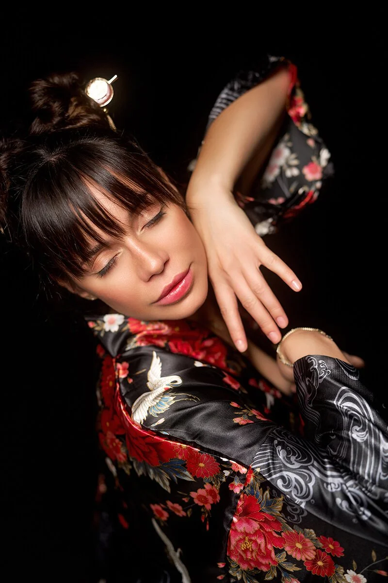 A woman with closed eyes lying on her side against a black background, wearing a black floral kimono, with her arm resting on her neck, a wrist bracelet, and a hair stick.