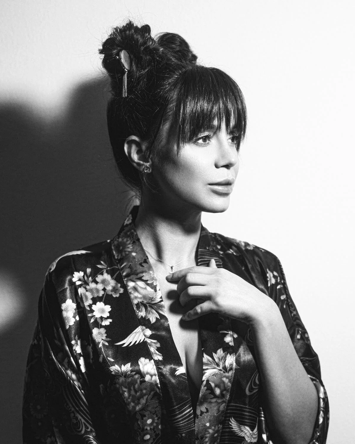 Black-and-white portrait of a woman with dark hair styled in an updo, wearing a floral patterned silk robe, jewelry, and gazing to the side with her hand near her collar.