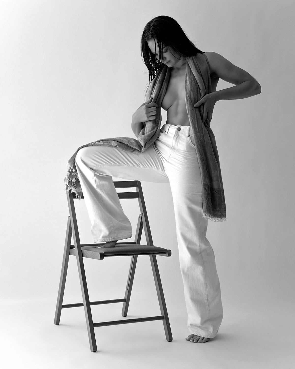 Black and white photo of a woman with long dark hair, topless, wearing loose white pants, standing with one foot on a chair, holding a scarf around her shoulders, against a plain light background.