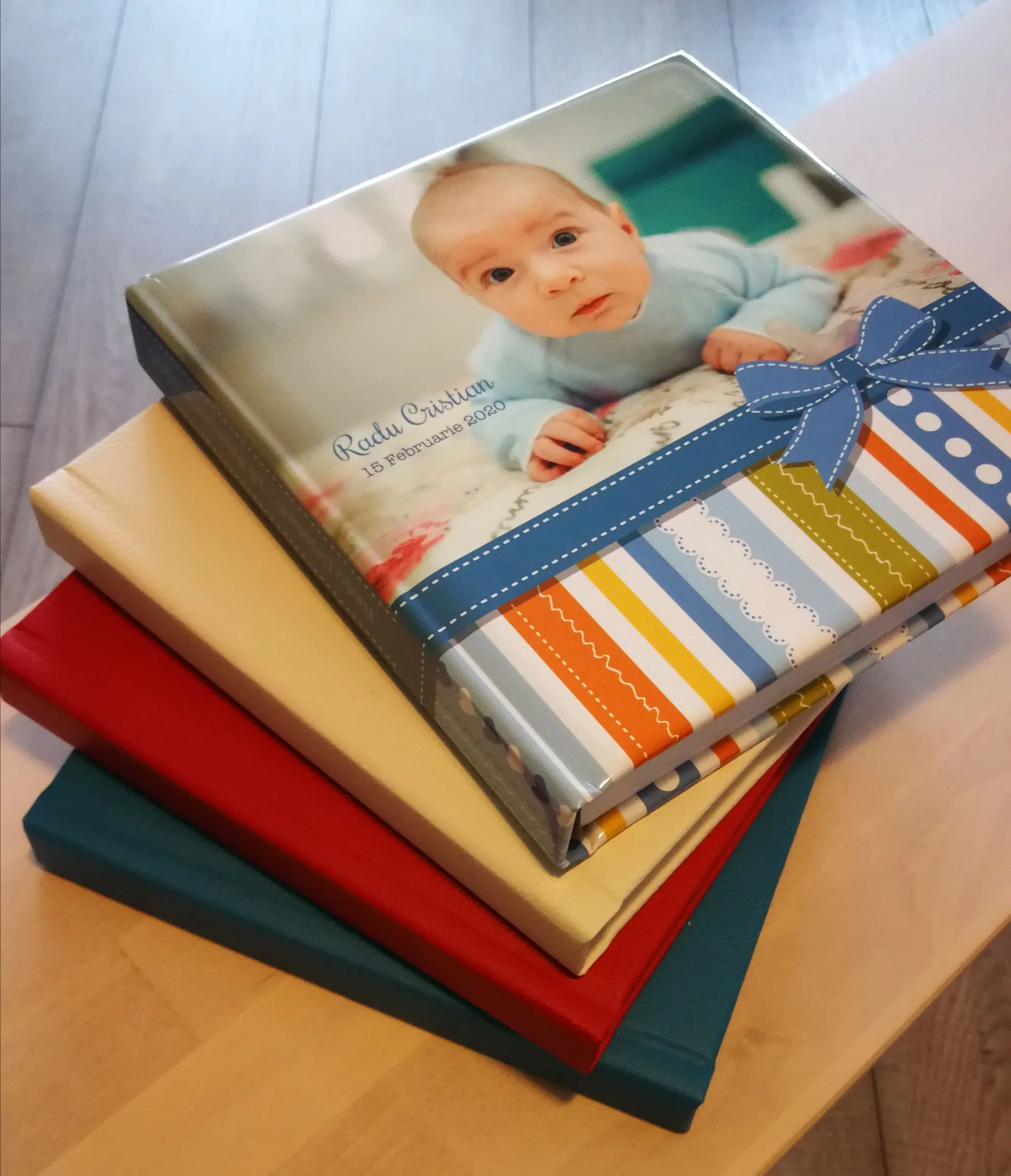 Stack of three photo albums on a wooden table, with the top album featuring a cover photo of a baby boy and the text 'Radu Cristian 15 Februarie 2020'.