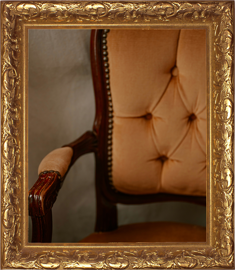 Born Again Consignment Parlor. Printers Row. Close-up of an elegant antique chair with a carved wooden frame, beige tufted upholstery, and brass nailhead trim, viewed through a decorative gold frame.