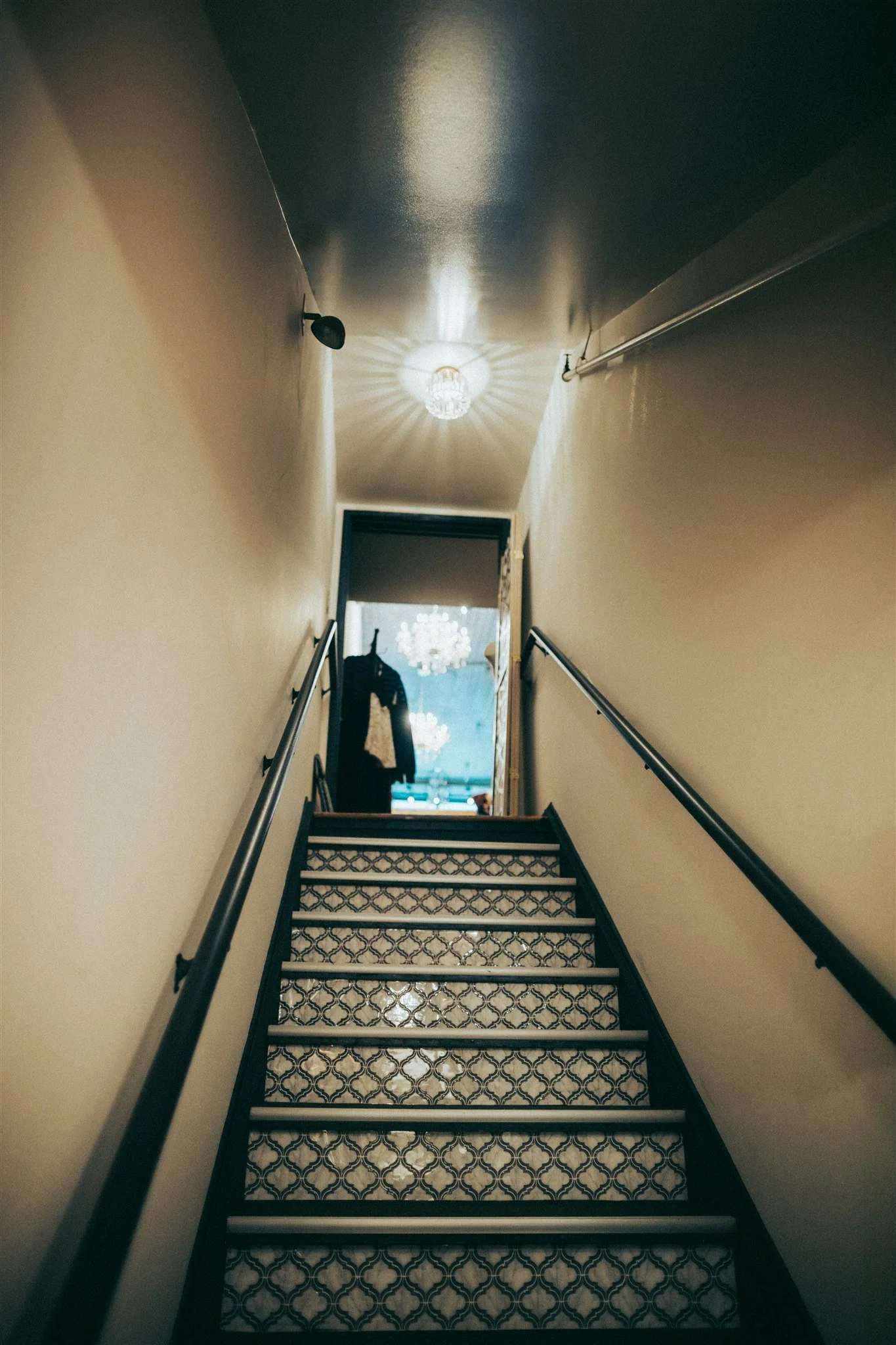 Born Again Consignment Parlor. Printers Row. Indoor staircase with patterned steps, beige walls, black handrails on both sides, a small ceiling light fixture, and an open door,
