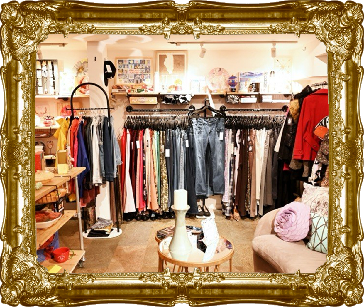 A boutique clothing store with racks of various clothing items, including pants, shirts, and jackets, on display. There is a small table with decorative items and a beige couch with pillows on the right side. Shelves with accessories and decor are in the background, surrounded by an ornate gold picture frame.