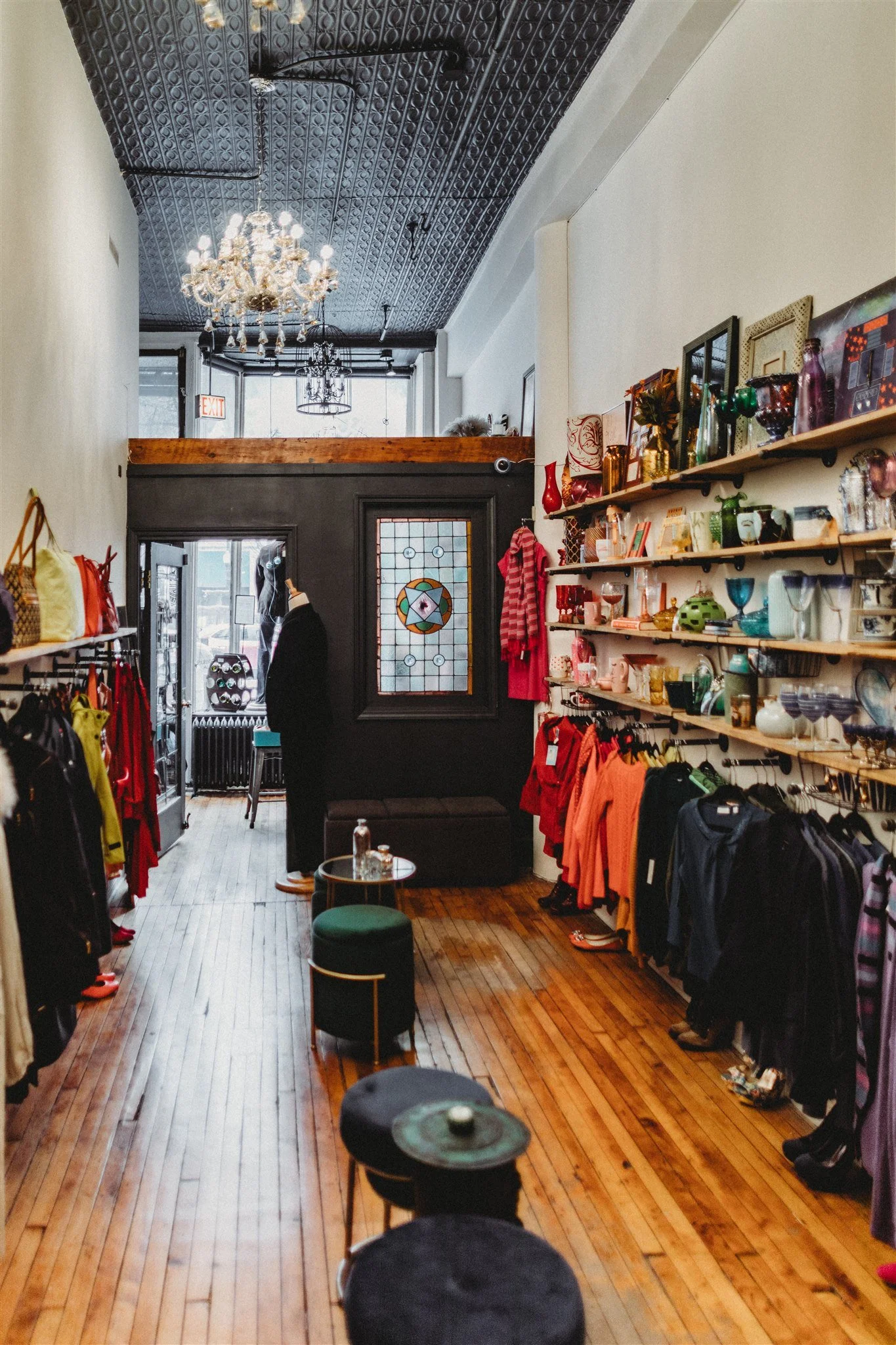 Born Again Consignment Parlor. Printers Row. A boutique store interior with clothing racks on both sides, shelves with decorative glassware and artwork, a vintage stained glass window, a chandelier, and a small seating area in the front.