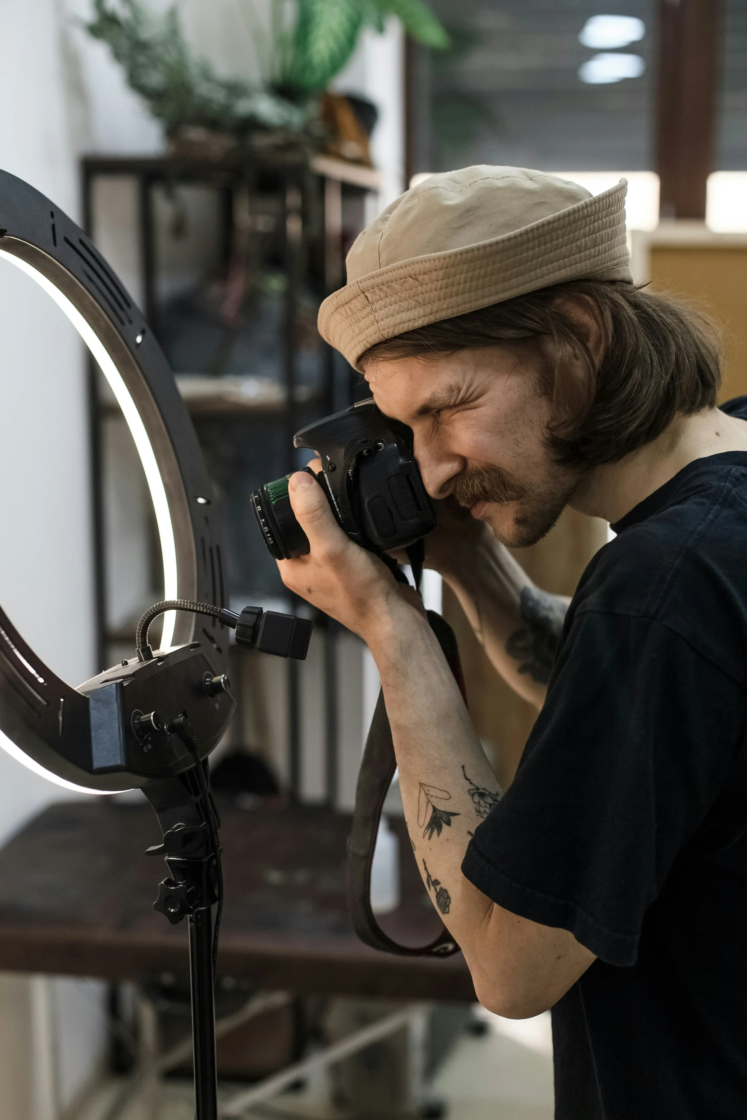 A man with a beige hat and tattoos on his arm is looking through a camera lens in a photography studio with a ring light.