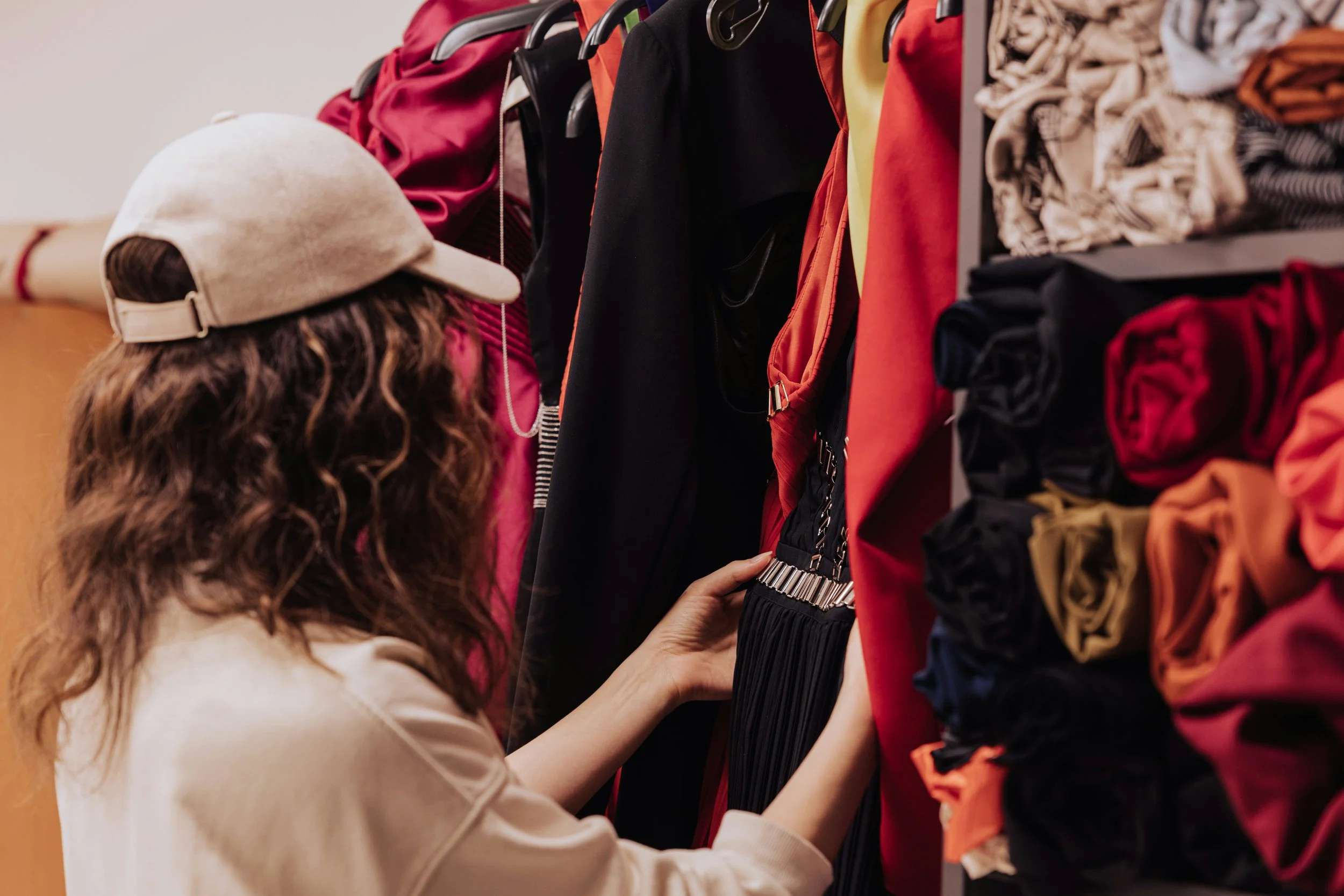 A woman with curly hair wearing a beige cap and a light-colored sweatshirt shopping for clothes, looking at hanging dresses in a store.