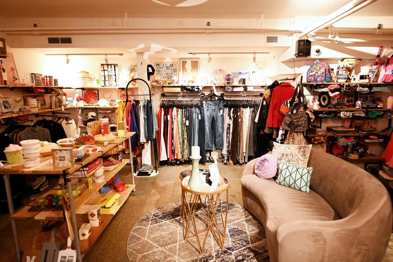 Born Again Consignment Parlor. Printers Row. A cozy boutique store with clothing on racks, shelves with accessories, and a sofa with decorative cushions, featuring warm lighting and eclectic decor.