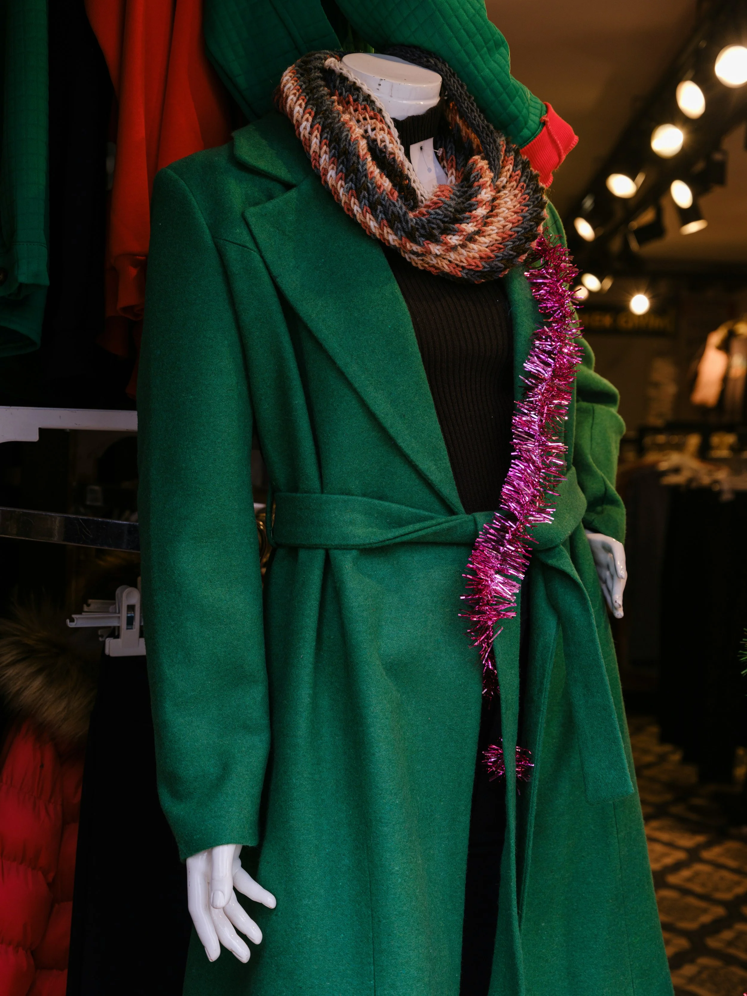 A mannequin dressed in a green coat, a colorful scarf, a black top, with pink tinsel hanging on the coat, in a clothing store.