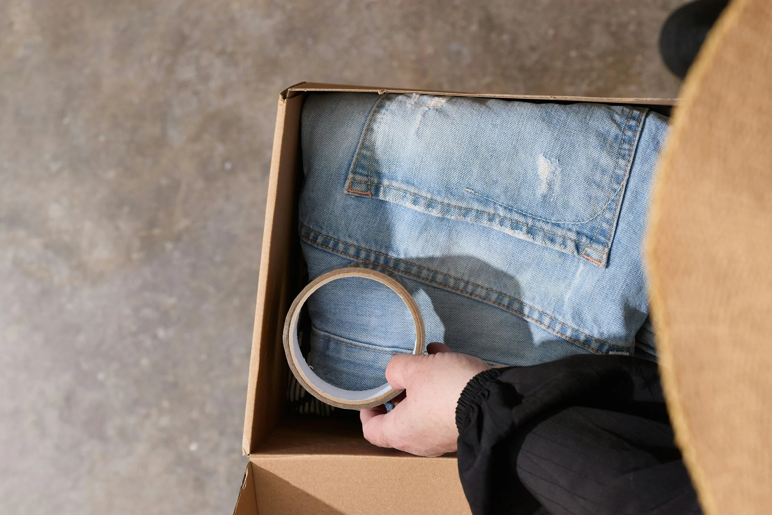 Person's hand holding a roll of packing tape inside a box with blue jeans.
