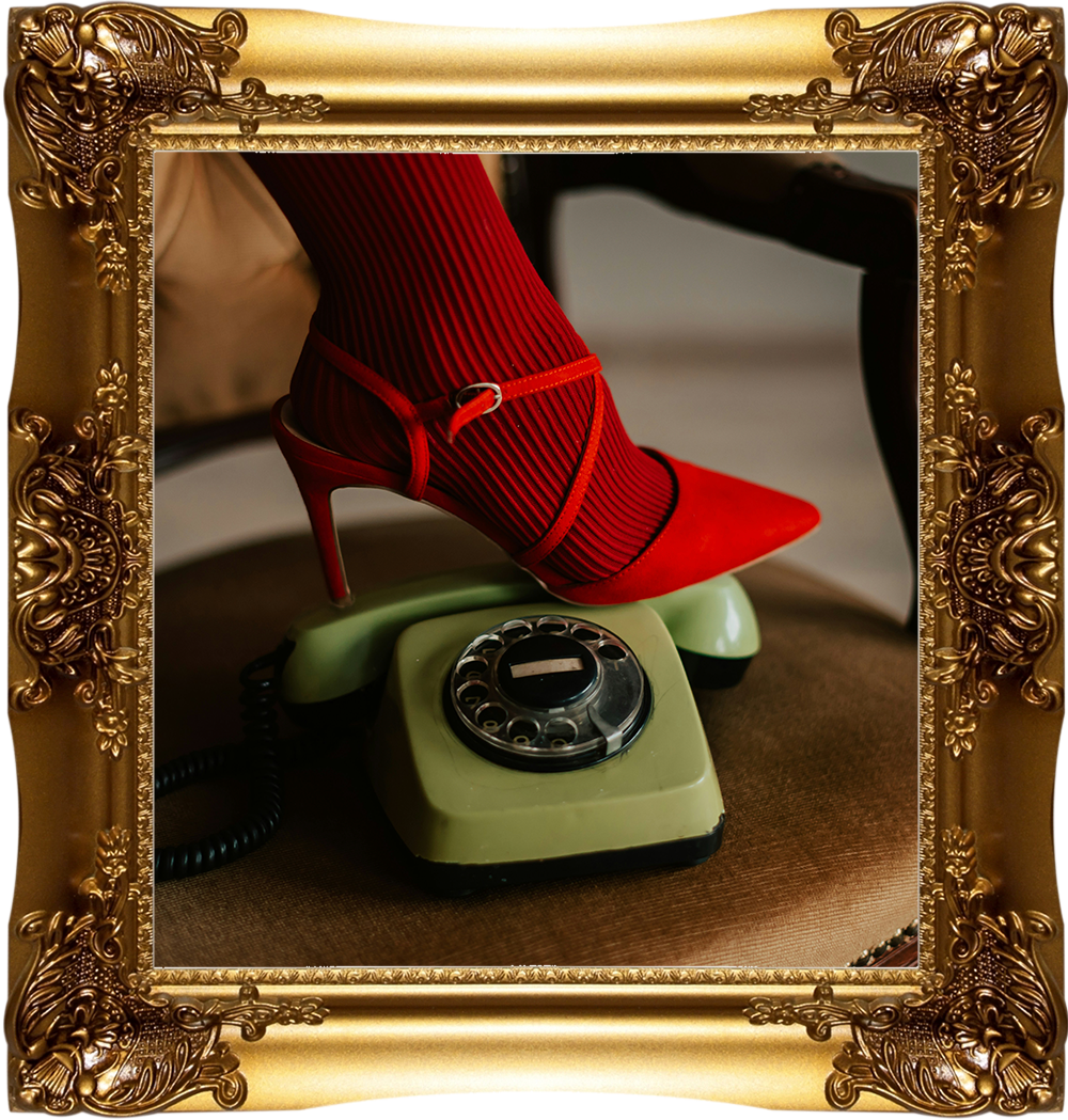 Born Again Consignment Parlor. Printers Row. A vintage green rotary telephone with a red high-heeled shoe and red stocking on the receiver, placed on a brown surface, framed with an ornate gold frame.