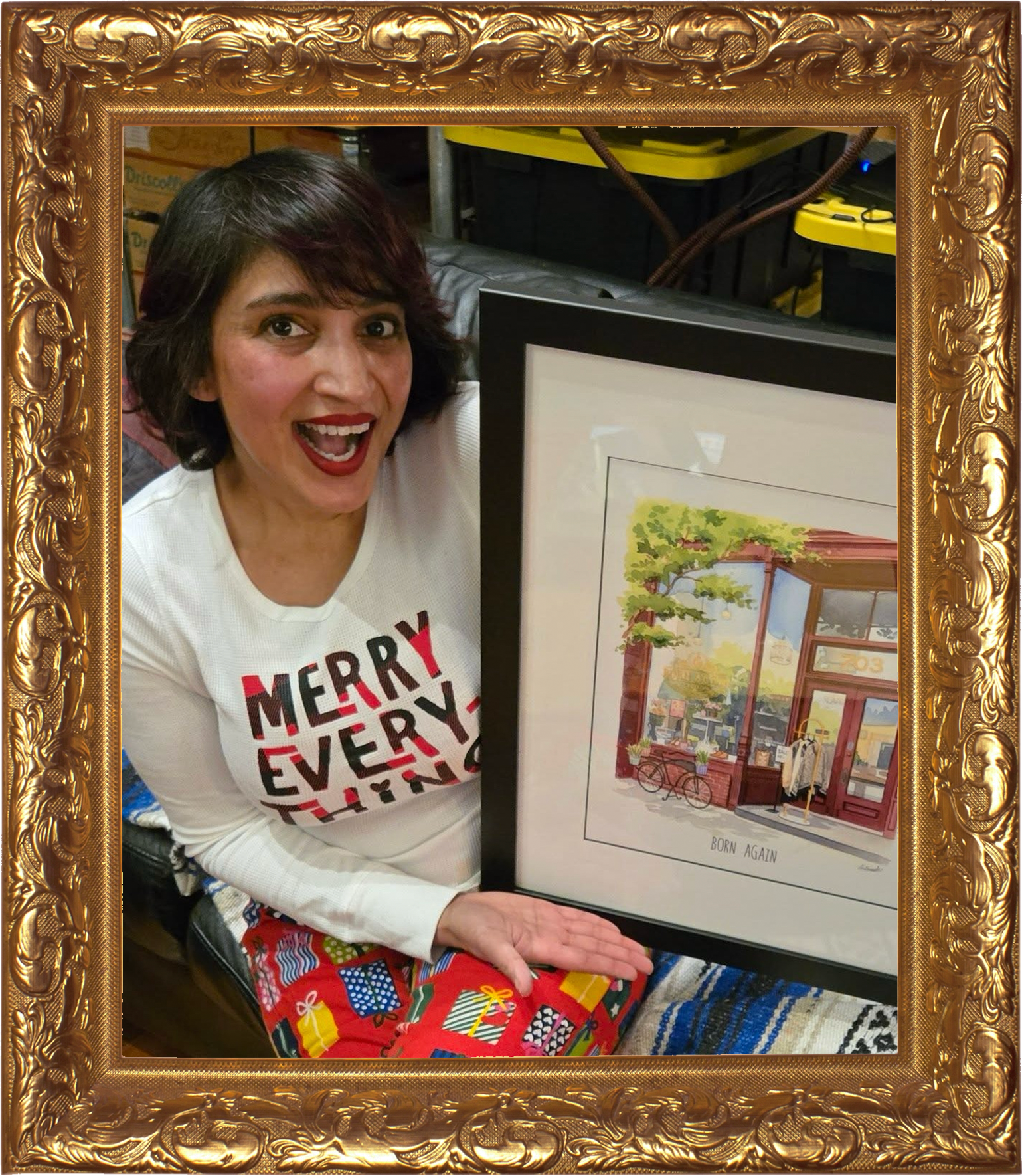 Summer Amin, wearing a white shirt with 'Merry Every' written on it and red patterned pants, is smiling and holding a framed artwork. The artwork depicts a storefront with large windows, a bicycle outside, and a tree with green foliage.