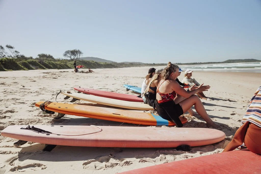 People sitting on surfboards on a sandy beach, talking and relaxing in sunny weather with the ocean waves in the background.