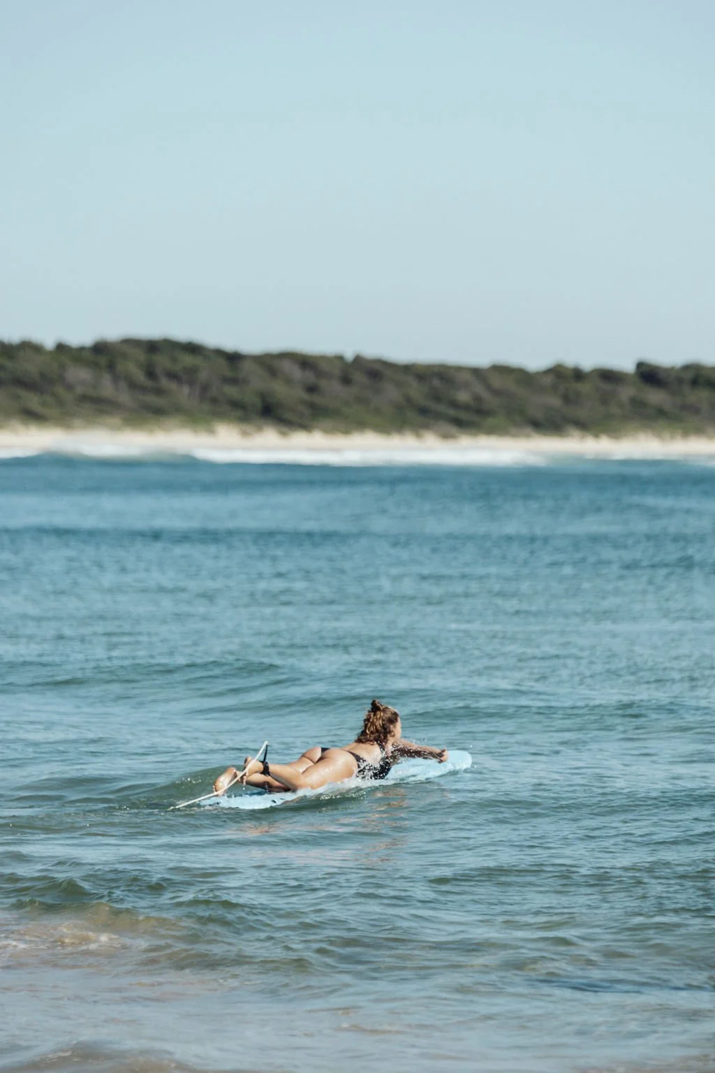 A woman lying on a surfboard paddling in the ocean near a sandy beach with green hills in the background.