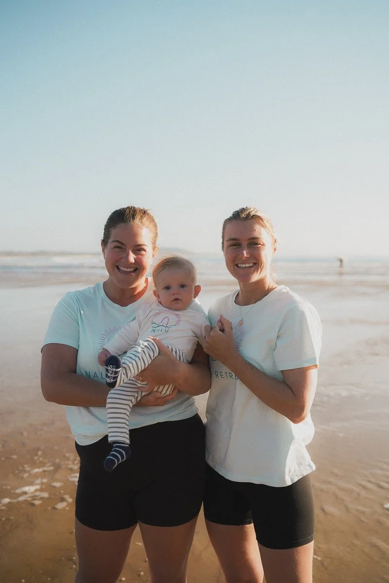 Two women standing on a beach holding a baby, all smiling at the camera, with the ocean and sky in the background.