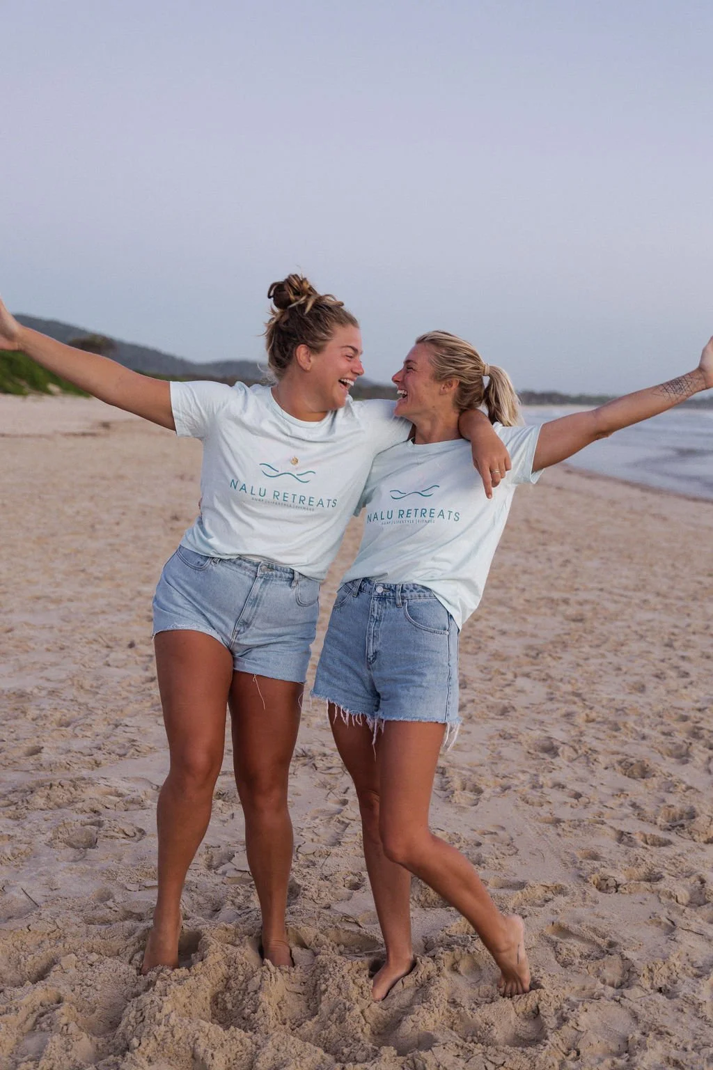 Two women smiling and celebrating on the beach, with arms around each other, wearing matching white t-shirts with "Nalu Retreats" logo, and denim shorts, near the water during sunset.