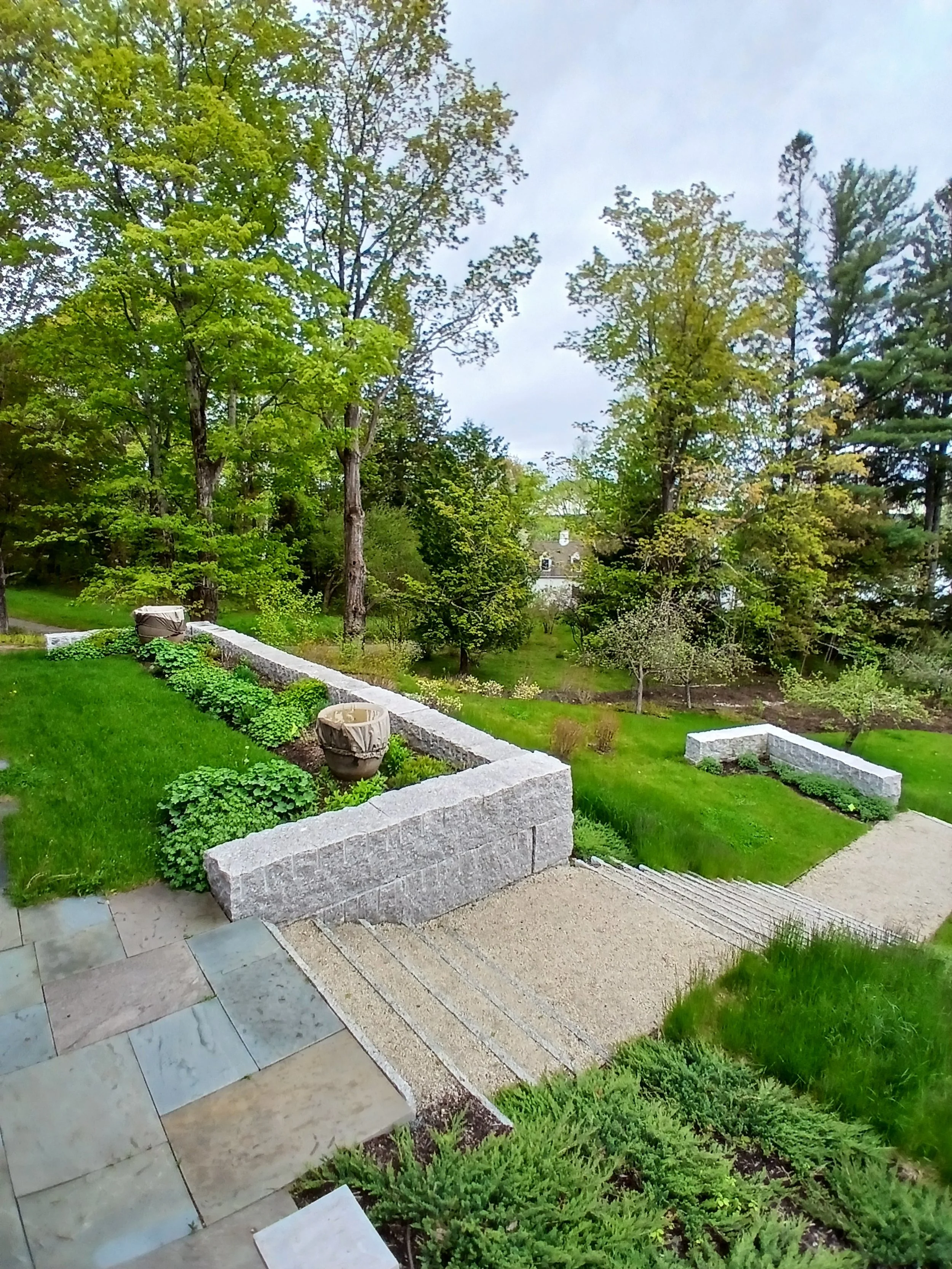A landscaped backyard with a stone staircase, green grass, trees, and flower beds, viewed from a patio or deck.