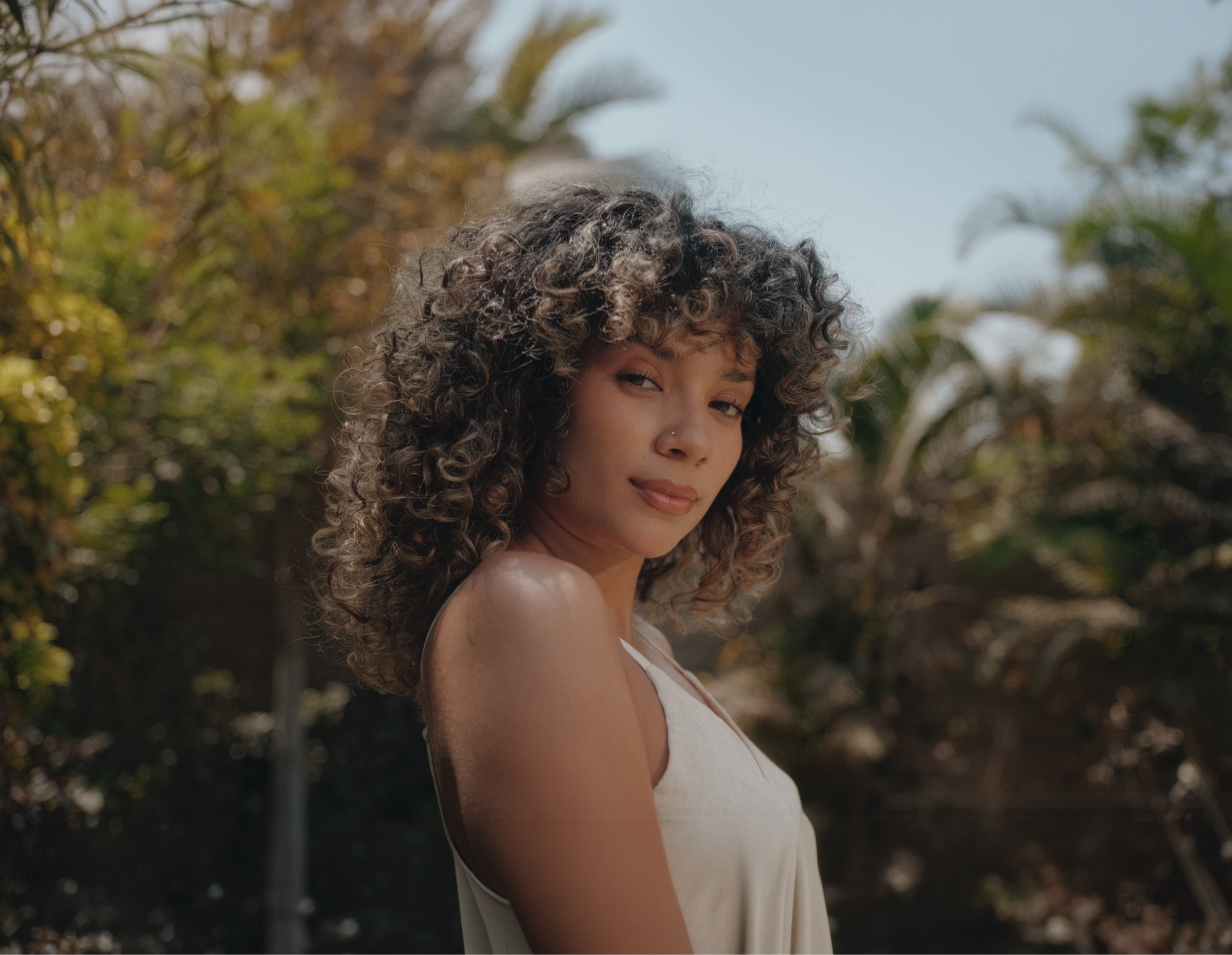 Mujer con rizos largos y cabello rizado, de piel clara, veste un vestido blanco y está al aire libre con fondo de árboles y cielo azul.