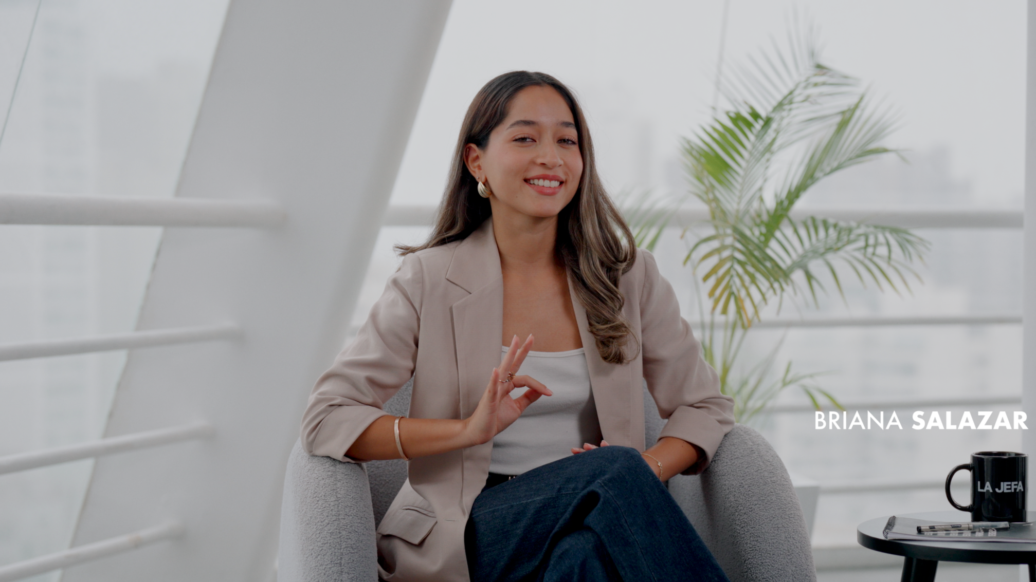 Mujer joven en una oficina moderna, sonriendo y haciendo una señal de paz con la mano, sentada en una silla gris. Tiene cabello largo y oscuro, usa blazer beige y camiseta blanca. Fondo con ventana grande y plantas verdes. Texto en esquina inferior d