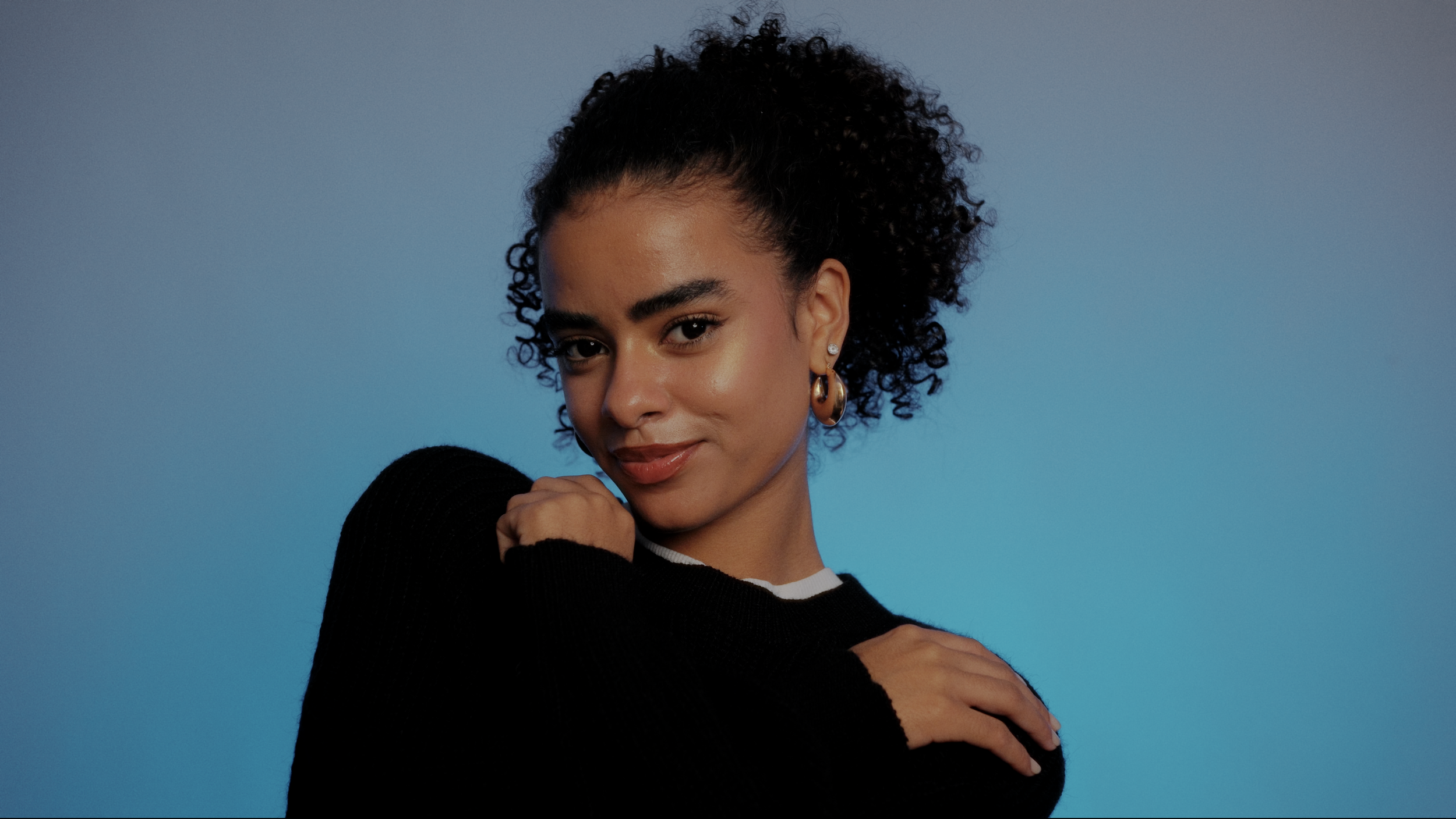 Joven mujer con cabello rizado y corto, usando aretes grandes y una camiseta blanca, con suéter negro, sonriendo y posando frente a un fondo azul.
