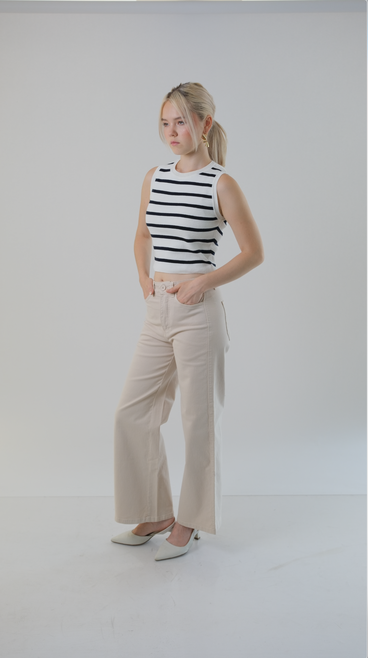 Joven con cabello rubio, puesto con blusa sin mangas de rayas blancas y negras, pantalones beige anchos y zapatos de tacón blanco, posando en un fondo blanco.