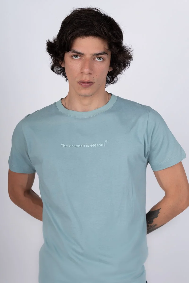 Joven con cabello oscuro y rubio, camiseta azul claro con el texto "La esencia es eterna", mirando fijamente a la cámara, en un fondo gris claro.